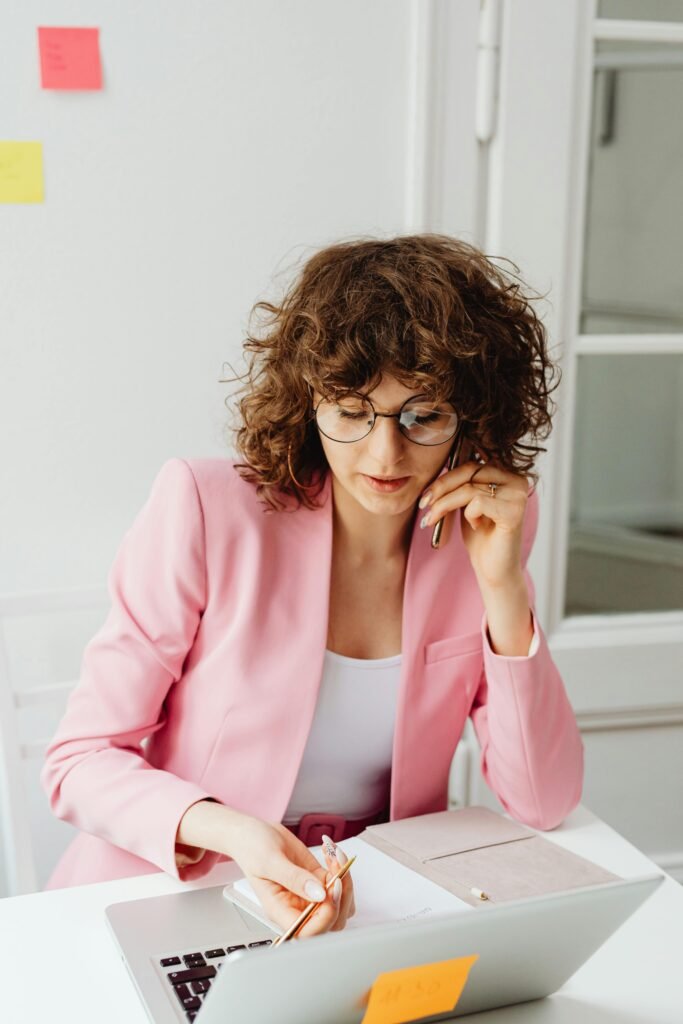 Focused woman in pink blazer working on laptop and phone.